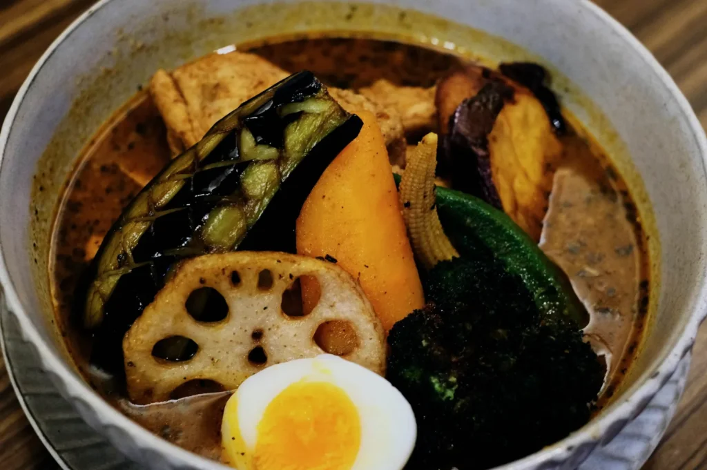 A bowl of Japanese curry with vibrant vegetables, including grilled eggplant, carrot, lotus root, sliced egg, broccoli, and baby corn, creating a hearty and rich tone.
