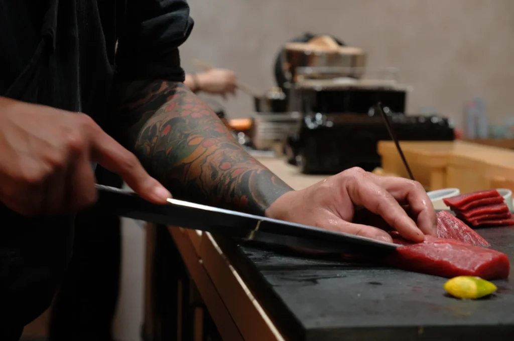 A tattooed Chef Masa expertly slices raw tuna on a dark counter, with kitchen tools blurred in the background. The scene conveys focus and craftsmanship.