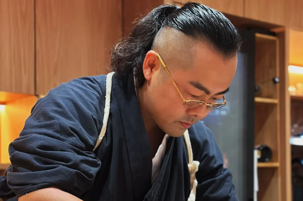Person with glasses and a modern hairstyle, focused on crafting something. They wear a dark robe in a warmly lit, wooden-paneled room, indicating a calm, attentive mood.