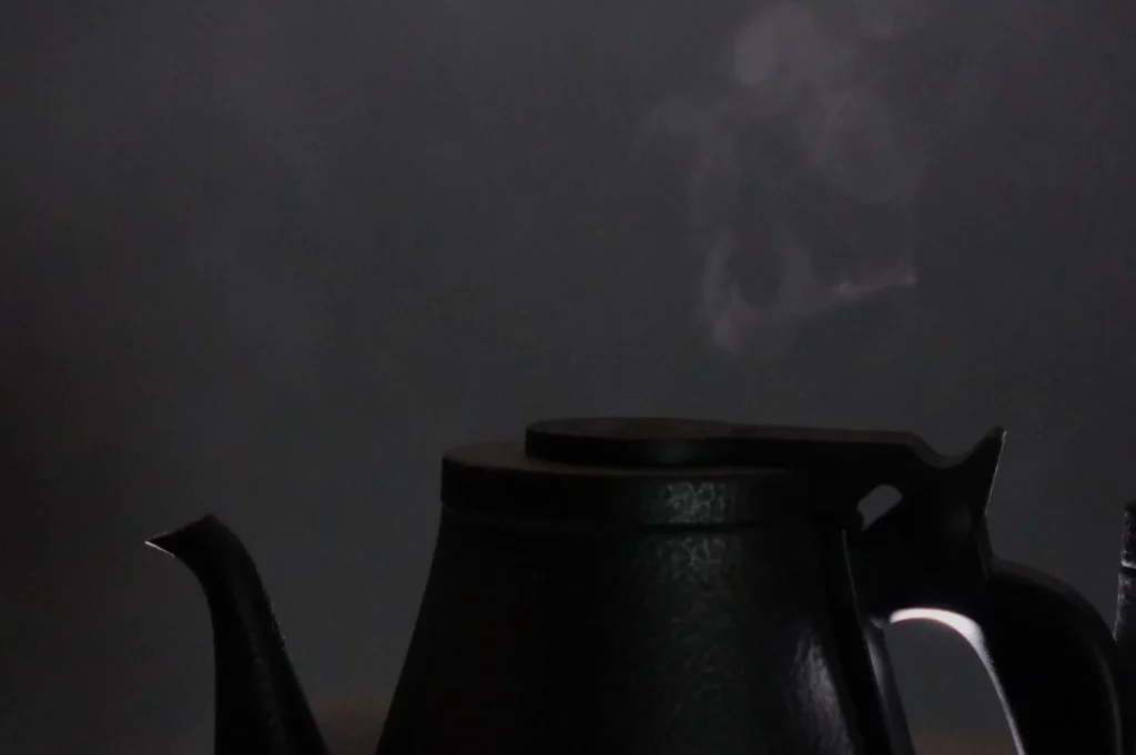 A black metal teapot with steam rising from the spout against a dark, blurred background. The setting is calm and minimalist, suggesting warmth.