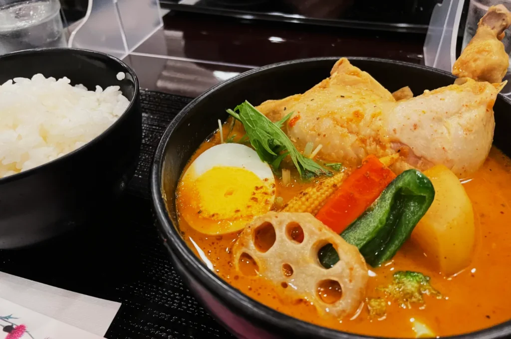 A colorful bowl of Japanese curry soup with a chicken leg, boiled egg, and assorted vegetables next to a bowl of steamed white rice. Cozy and appetizing.