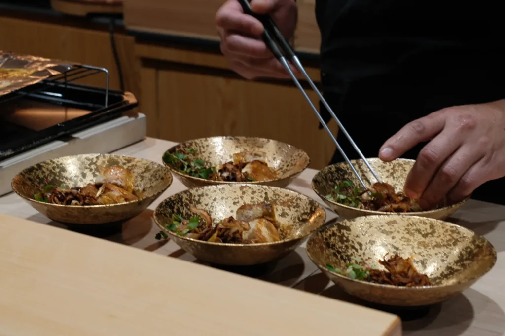 Chef Masa arranges food in six golden bowls using tongs. The bowls contain a gourmet dish with herbs, emphasizing precision and elegance in preparation.