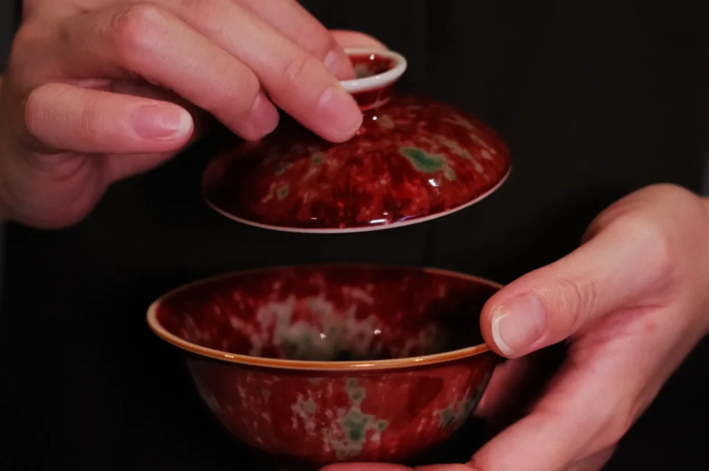 Hands delicately holding a patterned red and green ceramic bowl with a matching lid, suggesting careful handling and appreciation of craftsmanship.