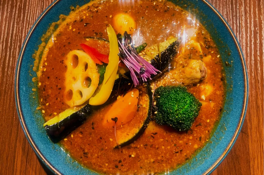 A vibrant bowl of soup curry with assorted vegetables including broccoli, lotus root, and peppers, served in a blue bowl on a wooden table.