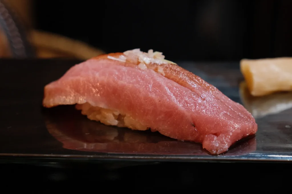 A piece of sushi featuring a slice of pink tuna over rice with a touch of garnish on top, resting on a dark plate. The background is softly blurred.