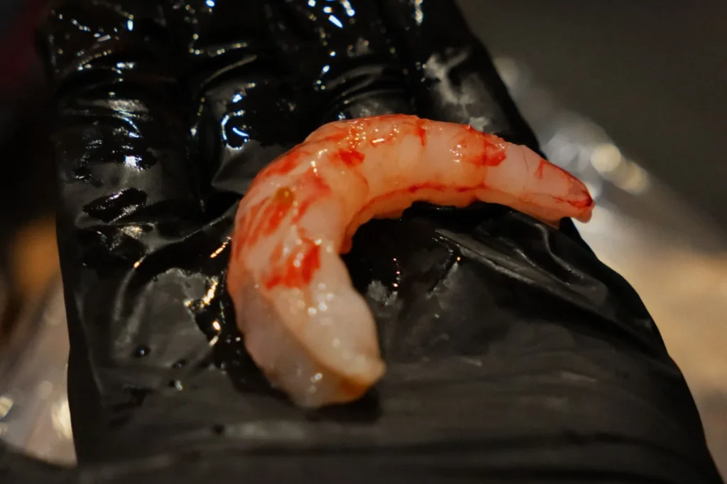 A close-up of a single raw shrimp with pink stripes placed on a glossy black glove. The background is blurred, emphasizing the shrimp's detail and texture.