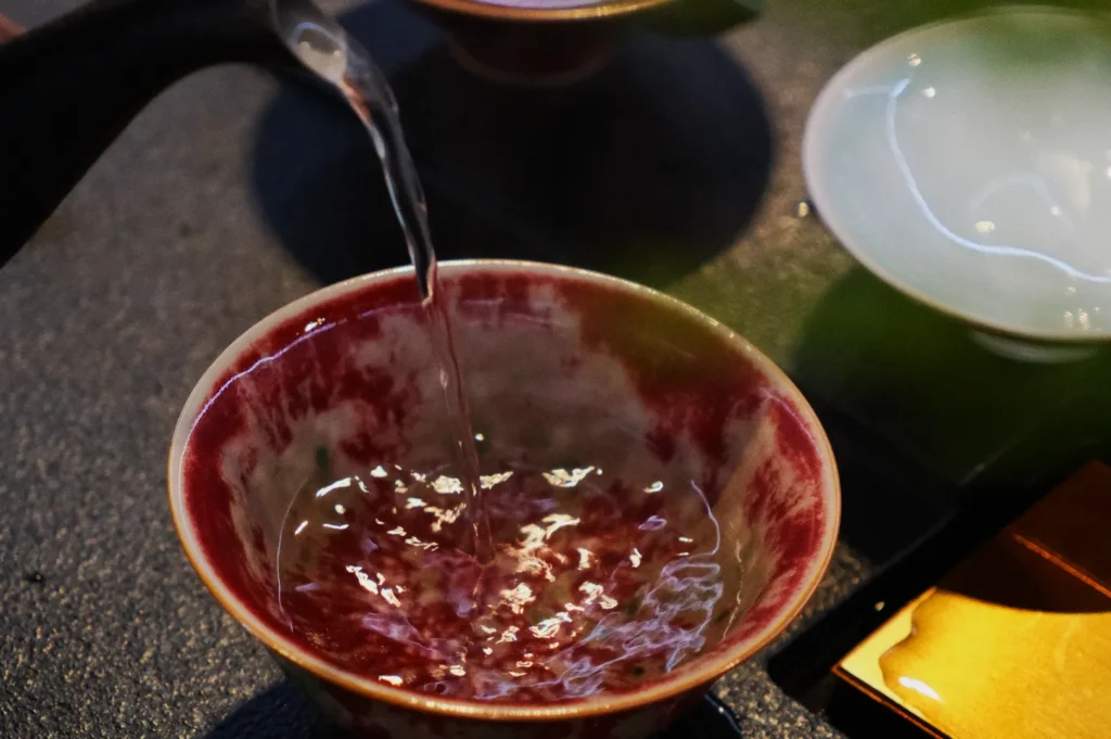 A patterned ceramic bowl with red and white swirls is being filled with clear liquid, possibly tea. Nearby are other cups and a warm, inviting atmosphere.