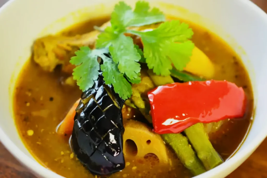 A bowl of colorful curry soup with vibrant veggies like eggplant, red pepper, asparagus, lotus root, and garnished with cilantro leaves.