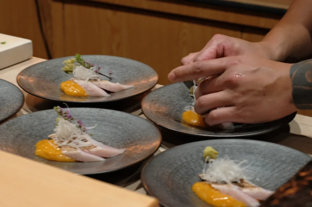 Chef Masa’s hands carefully plating artful sushi with white fish, mango puree, and garnishes on gray dishes. The scene conveys precision and culinary artistry.