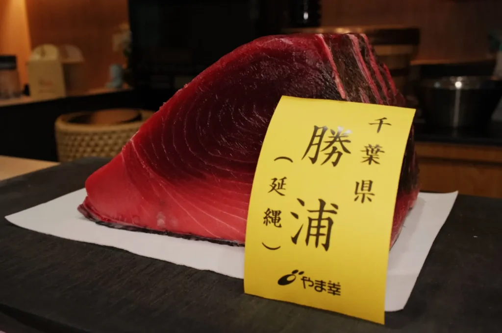 A large, fresh cut of tuna is displayed on a kitchen counter, partially wrapped in paper. A yellow label with Japanese text is attached to the fish.