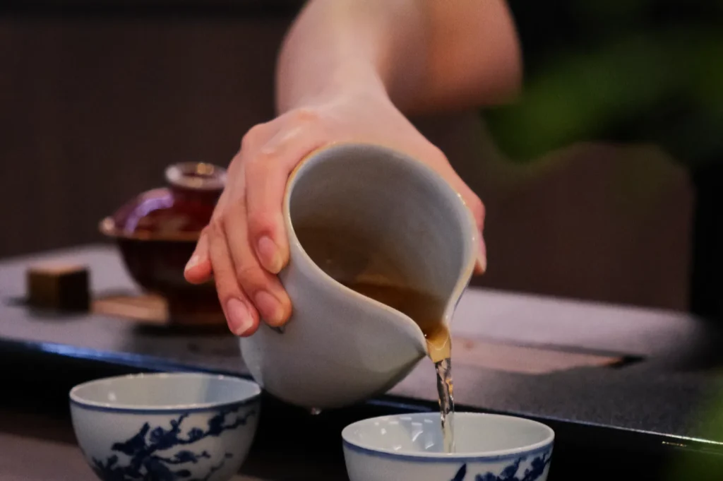 A hand gracefully pours tea from a ceramic pitcher into blue and white teacups on a tray, evoking a calm and traditional atmosphere.