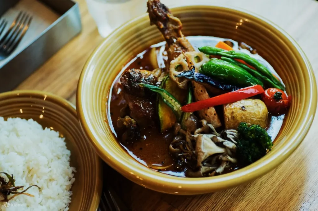 A bowl of flavorful curry with assorted vegetables and a chicken drumstick, served beside a dish of steamed white rice on a wooden table.