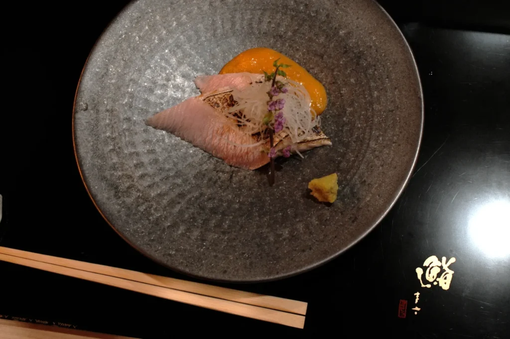 A stylish dish featuring thinly sliced sashimi garnished with white radish and purple flowers on a dark gray plate. Beside it are orange sauce and wasabi. Chopsticks and Japanese script are visible on the black surface, evoking elegance.