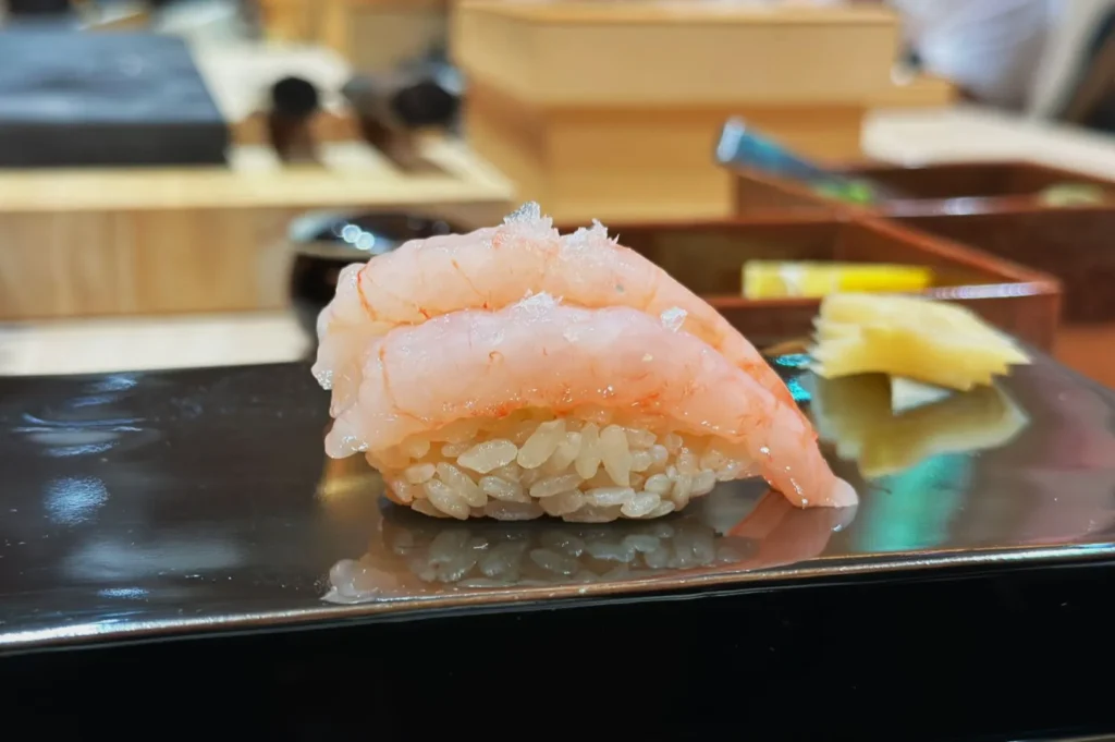 Close-up of a sushi piece with translucent shrimp on rice, garnished with salt. A side of pickled ginger is in the background on a sleek black plate.