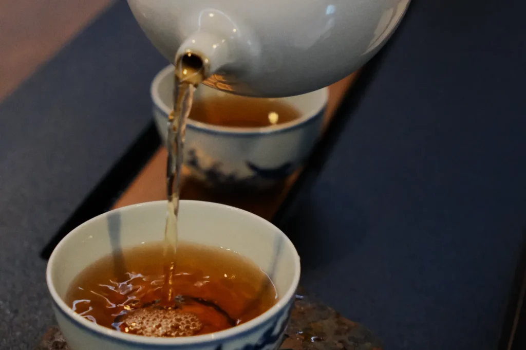 A white teapot pouring amber-colored tea into a delicate blue and white porcelain cup. The scene is calm and inviting, capturing the essence of a traditional tea ceremony.