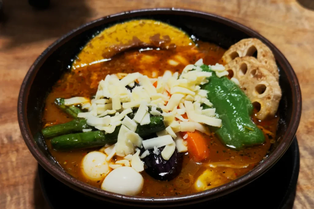 A rich soup in a dark bowl with vibrant vegetables, including green beans, peppers, and lotus root. Topped with shredded cheese, evoking warmth.