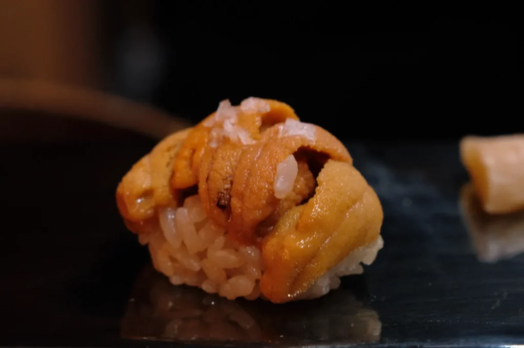 Close-up of a uni sushi piece on a dark surface, featuring creamy orange sea urchin atop vinegared rice. The setting is minimalist, evoking elegance.
