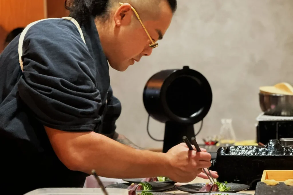 Chef carefully plating a dish in a modern kitchen, using tweezers for precision. The setting is professional, focused, and creative.
