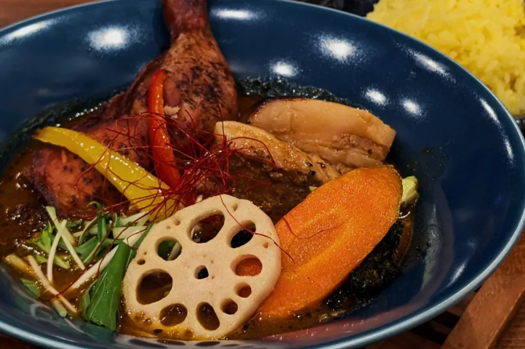 A vibrant dish featuring a roasted chicken leg, colorful bell peppers, carrots, and lotus root in a rich curry sauce, served in a blue bowl.