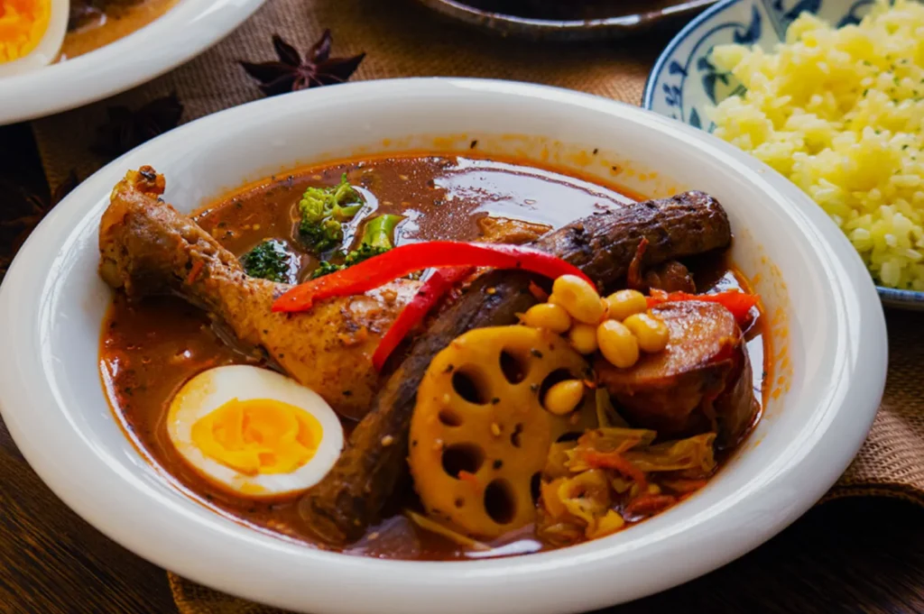 Bowl of spicy curry soup with chicken drumstick, boiled egg, lotus root, red pepper slices, and peanuts, garnished with broccoli. Served with a side of yellow rice.
