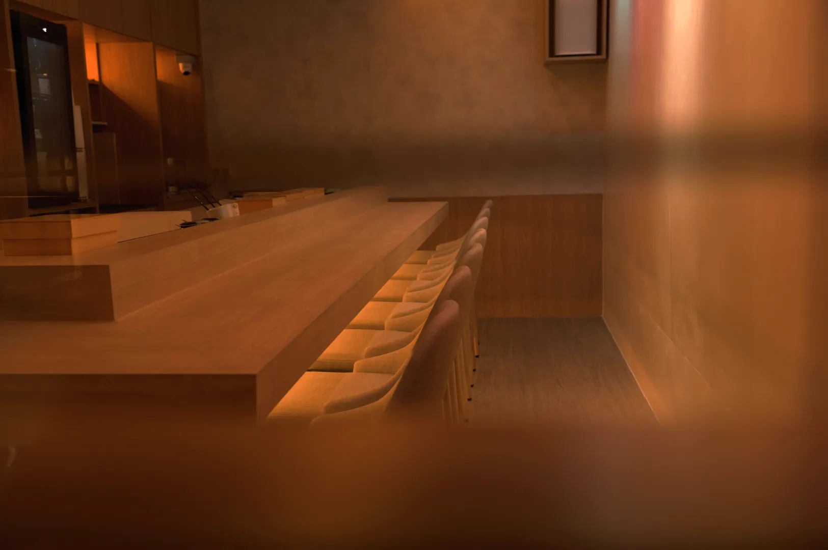 Warmly lit, minimalist interior of a dimly lit restaurant with a long wooden counter and neatly arranged wooden chairs. The ambiance is cozy and intimate.