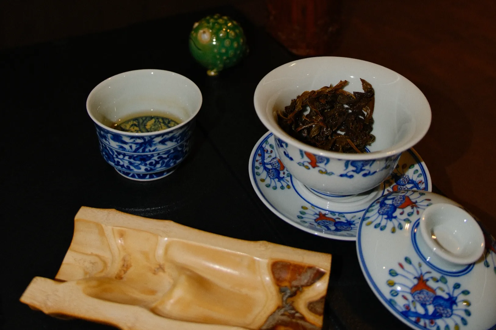 A traditional Asian tea set with blue floral designs is displayed. A gaiwan with tea leaves, a small cup with green tea, and a decorative green item are seen, creating a calm and cultural ambiance.