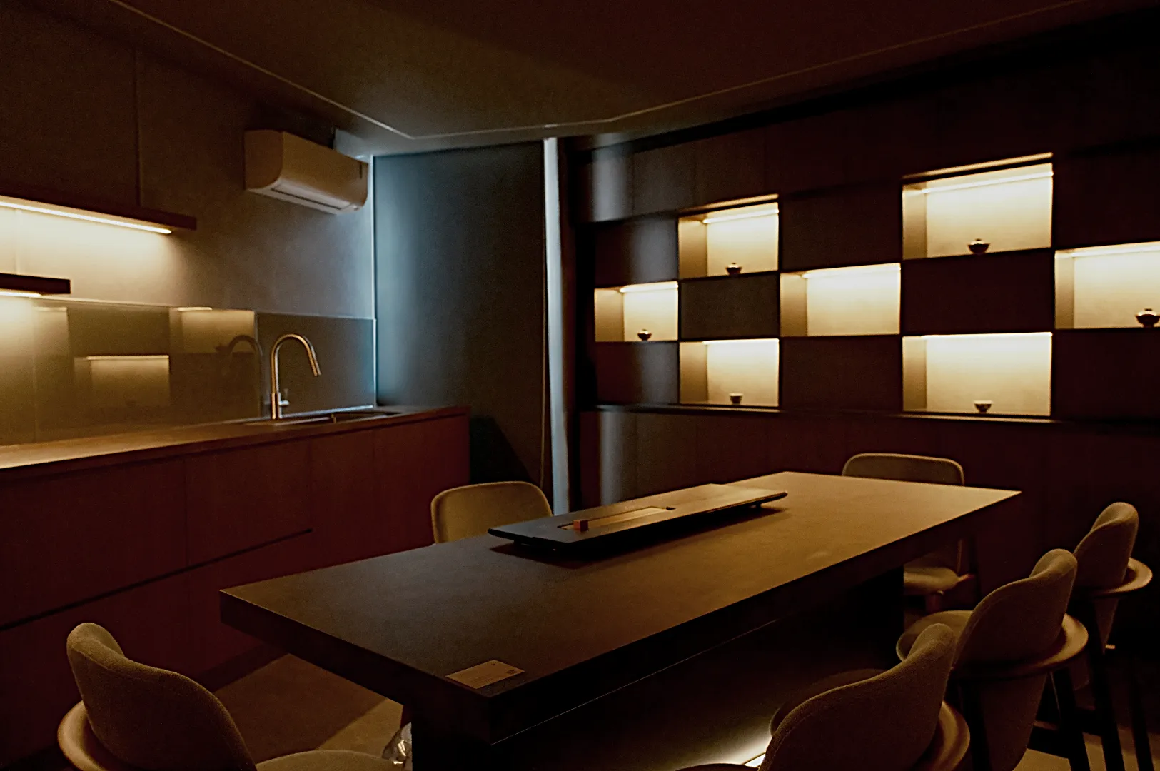 A modern, dimly lit dining room features a sleek wooden table with chairs. The wall has illuminated shelves displaying small vases, creating a cozy atmosphere.