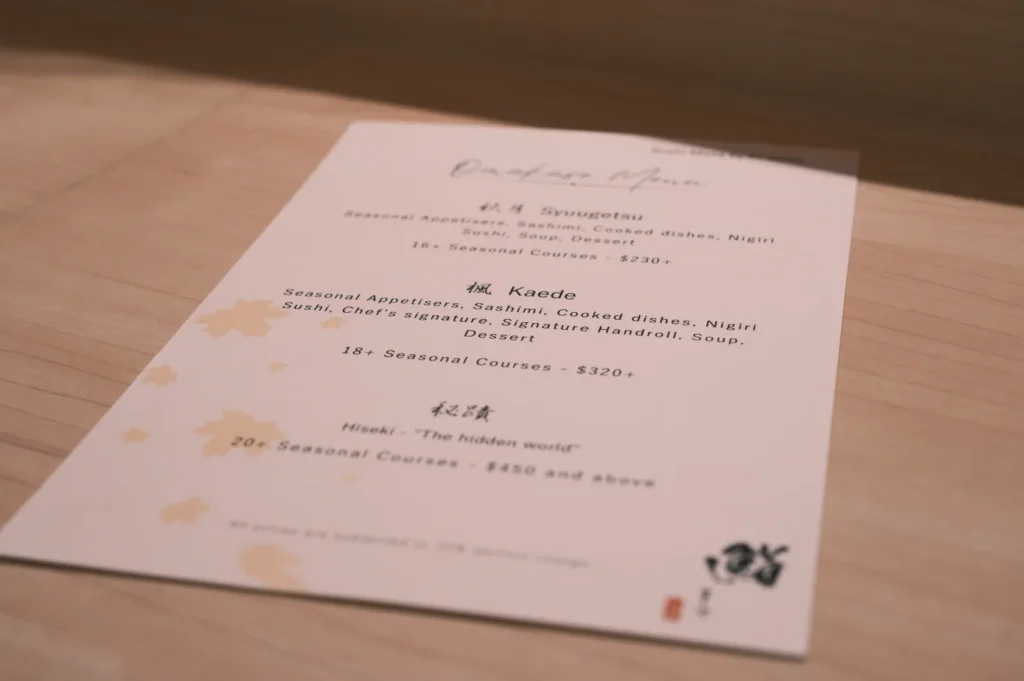 A tan menu titled "Dinner Menu" on a wooden table. It lists three Japanese course options with prices, featuring elegant maple leaf motifs.