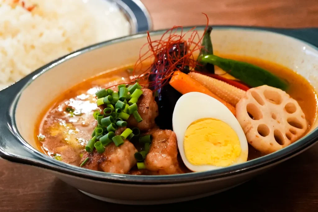 A bowl of colorful curry soup with chicken, topped with chopped green onions. Garnished with a halved boiled egg, lotus root slice, carrots, and chili peppers. Rice is partially visible in the background.