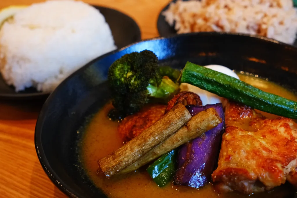 A bowl of food featuring a piece of grilled chicken, vegetables such as broccoli and green beans, and a rich sauce, accompanied by servings of rice on the side.