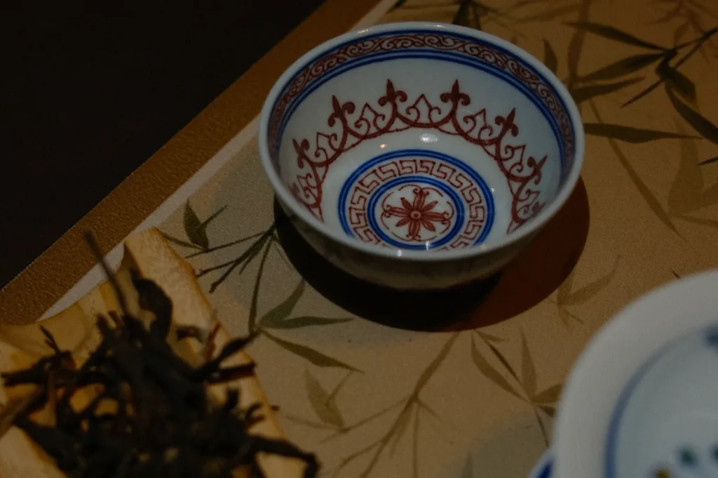 A ceramic bowl with intricate red and blue geometric patterns on the inside, placed on a bamboo-patterned mat. Nearby are dried tea leaves.