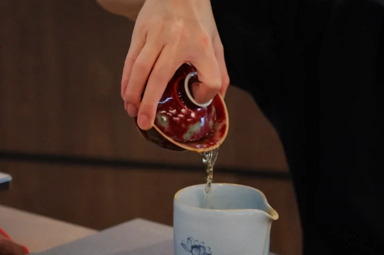 Close-up of a hand pouring tea from a small, red ceramic pot into a white cup with a floral design. The setting is calm and focused.