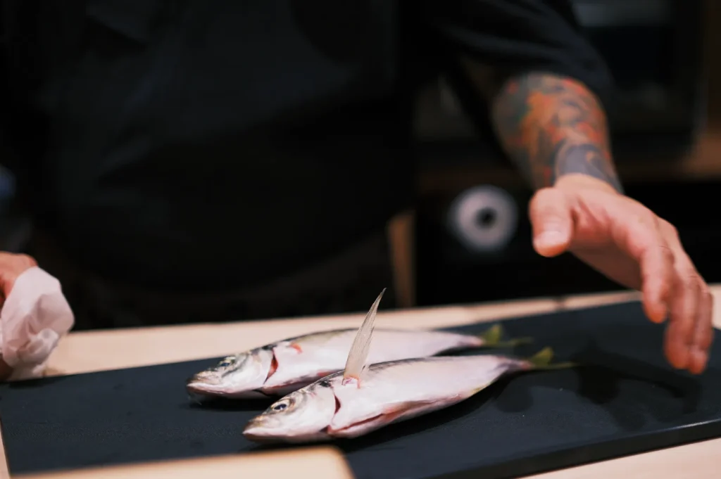 A chef with tattooed arms prepares two whole fish on a black cutting board. The setting suggests a professional kitchen environment, focused and meticulous.