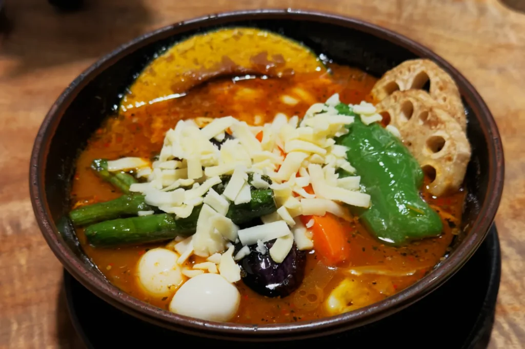 A bowl of vibrant vegetable soup topped with shredded cheese. Includes green peppers, quail eggs, lotus root, in rich broth, on a wooden table.
