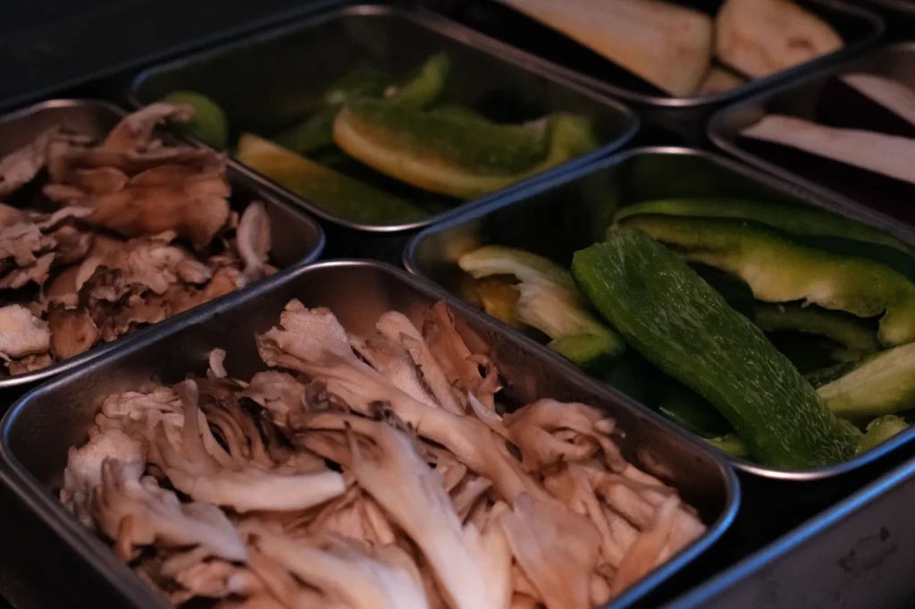 Assorted sliced vegetables including mushrooms, bell peppers, and zucchini in metal trays.