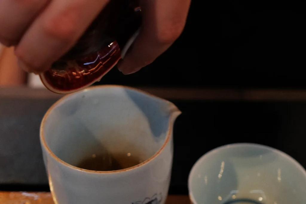 A hand pours tea into a small, white ceramic pitcher beside an empty teacup. The focus is on the action, with a calm and deliberate tone.