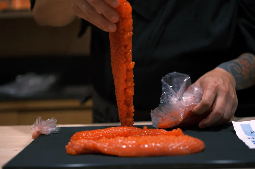 A person in dark clothing processes vibrant orange fish roe, creating a fresh and meticulous culinary scene focused on precision and craftsmanship.