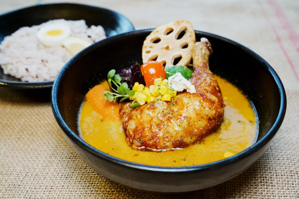 Delicious curry dish with a chicken leg, vegetables, and lotus root slice in a dark bowl. Side of rice and eggs in background, on burlap surface.