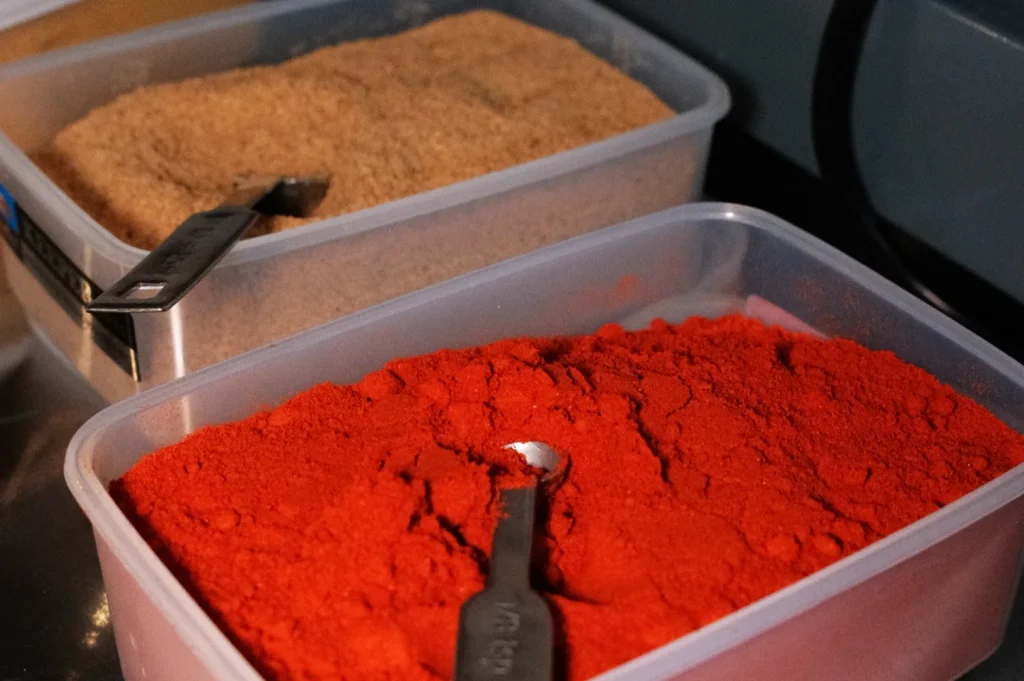 Two containers filled with different types of spices, one with a light brown powder and the other with a vibrant red powder, each with a measuring spoon placed inside.