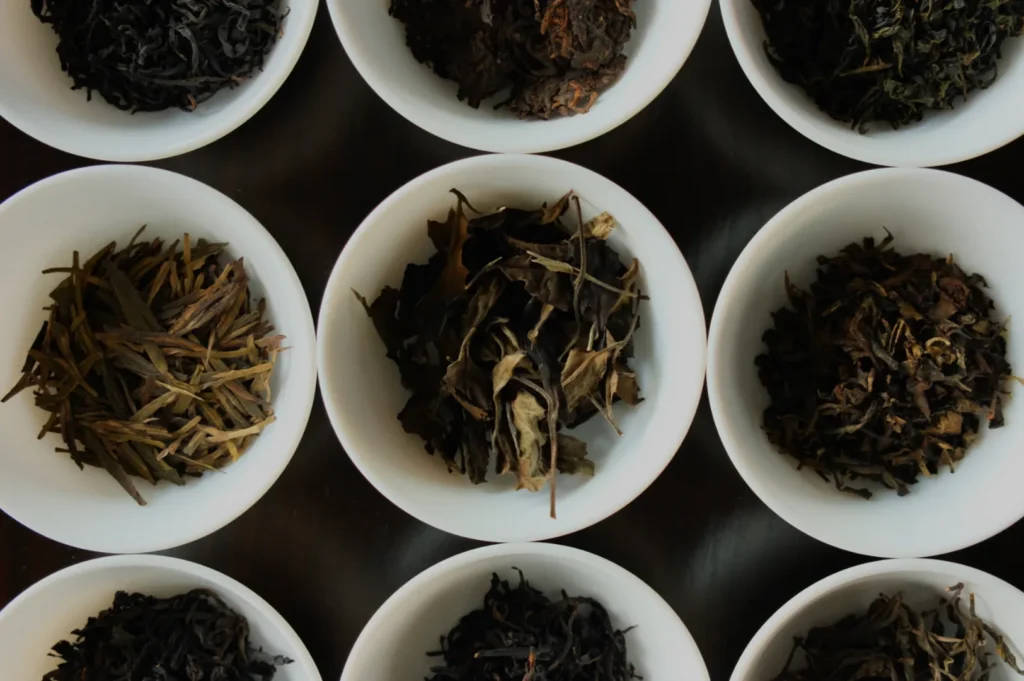 Bowls of various dried tea leaves are arranged in a grid on a dark surface. Each white bowl contains different tea varieties, showcasing diverse textures and colors.