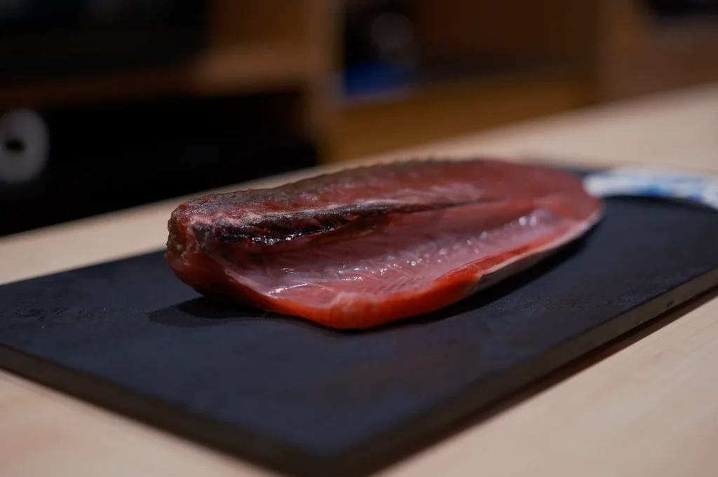 Close-up of a raw fish fillet, likely tuna, on a black cutting board, highlighting its rich red color and glossiness against a soft-focus background.