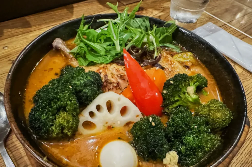 A vibrant bowl of soup features broccoli, red pepper, arugula, lotus root, and a quail egg. The rich broth exudes warmth and comfort.