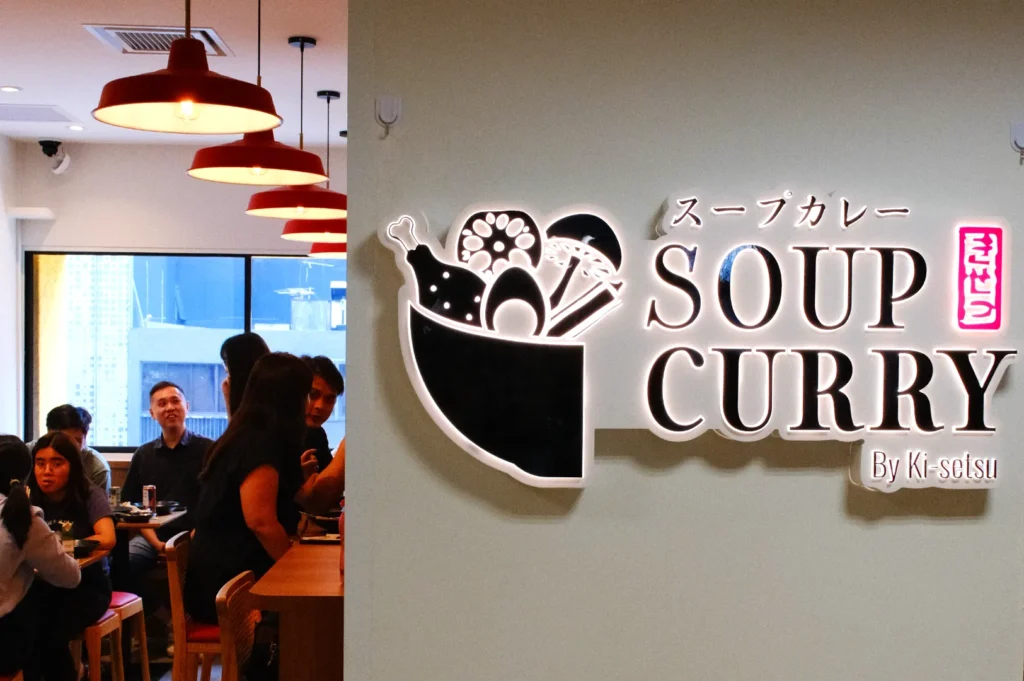 A restaurant interior with a sign that reads "SOUP CURRY" with Japanese text and a logo featuring food items.