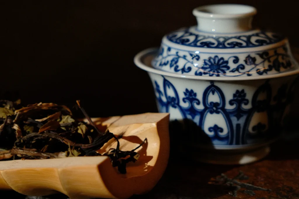 A decorative blue and white porcelain cup with a detailed floral pattern beside a wooden tray holding dried tea leaves. The scene exudes a warm, rustic charm.