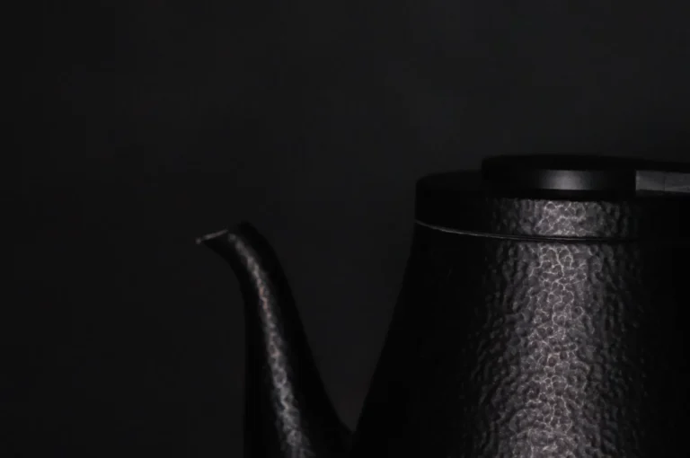 Close-up of a black, textured metal teapot against a dark background. The image focuses on the spout and upper body, conveying a minimalist, elegant tone.