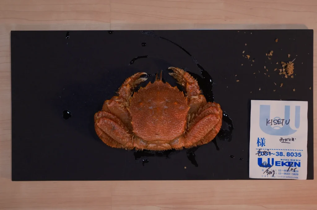 A cooked crab sits on a dark rectangular mat placed on a light wooden surface. Beside it is a folded receipt with Japanese text and a logo.