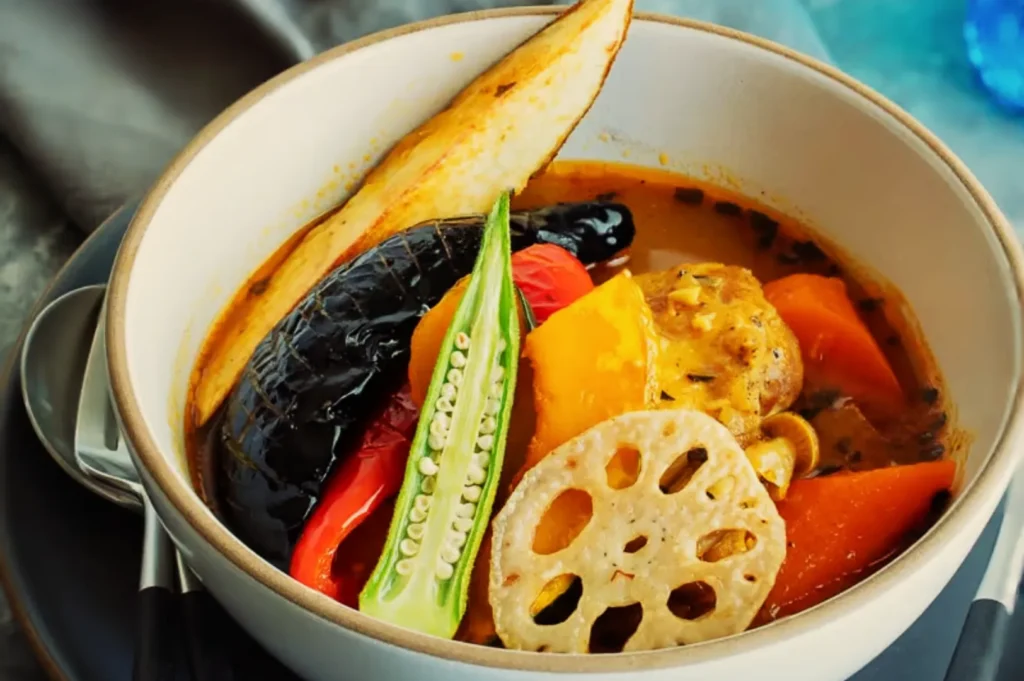 A bowl of colorful vegetable curry with eggplant, carrot, okra, lotus root, and a potato wedge in rich broth, exuding warmth and comfort.