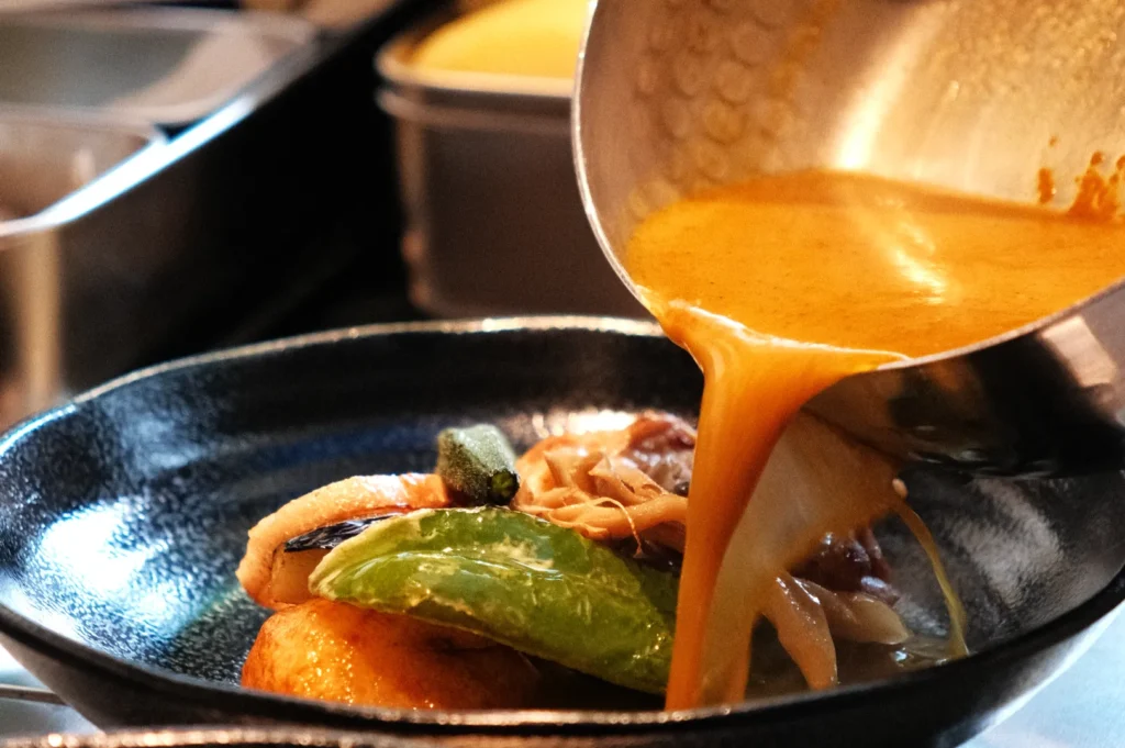 A dish being served with sauce being poured over it, featuring vegetables and a protein on a black plate.