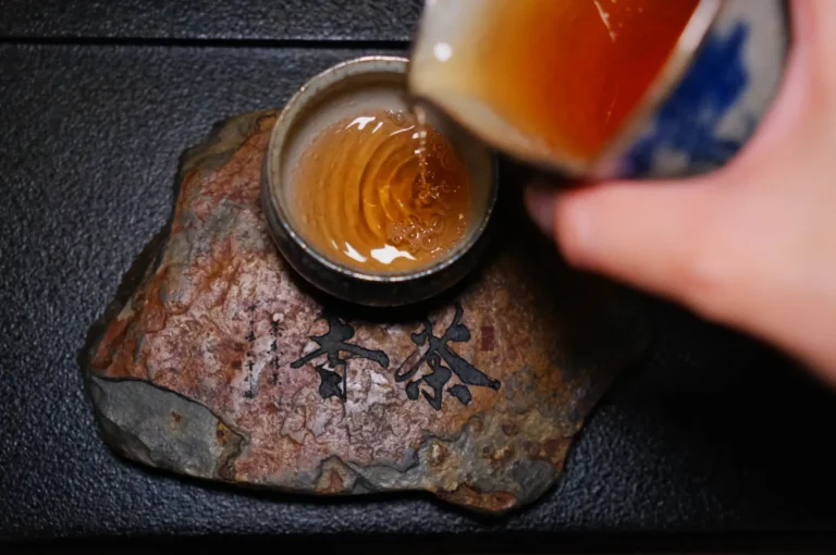 Hand pouring tea into a ceramic cup on a textured, stone-like surface with Asian characters. The scene conveys a traditional, serene vibe.
