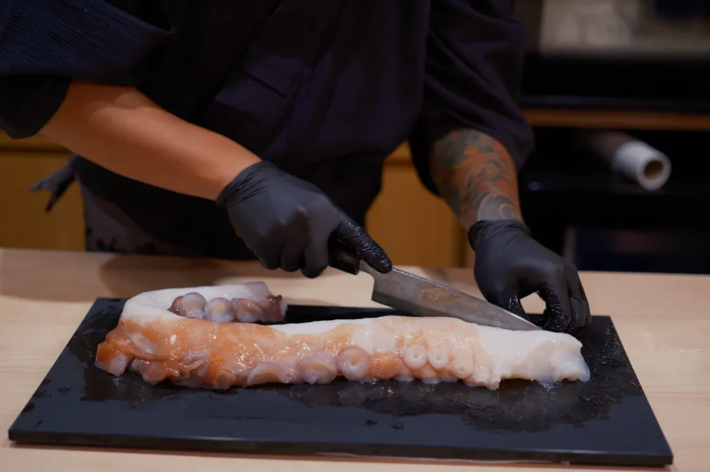 Chef Masa with tattooed arms and black gloves is slicing an octopus tentacle on a dark cutting board, conveying precision and culinary skill.